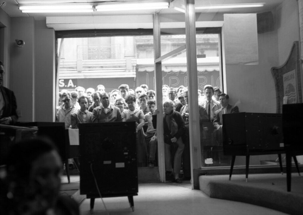 Personas asomadas al aparador de una tienda observan por televisión la visita del presidente de Estados Unidos John F. Kennedy a la Ciudad de México, junio de 1962. Colección Archivo Casasola ©19078 SC-INAH-SINAFO-Fototeca Nacional