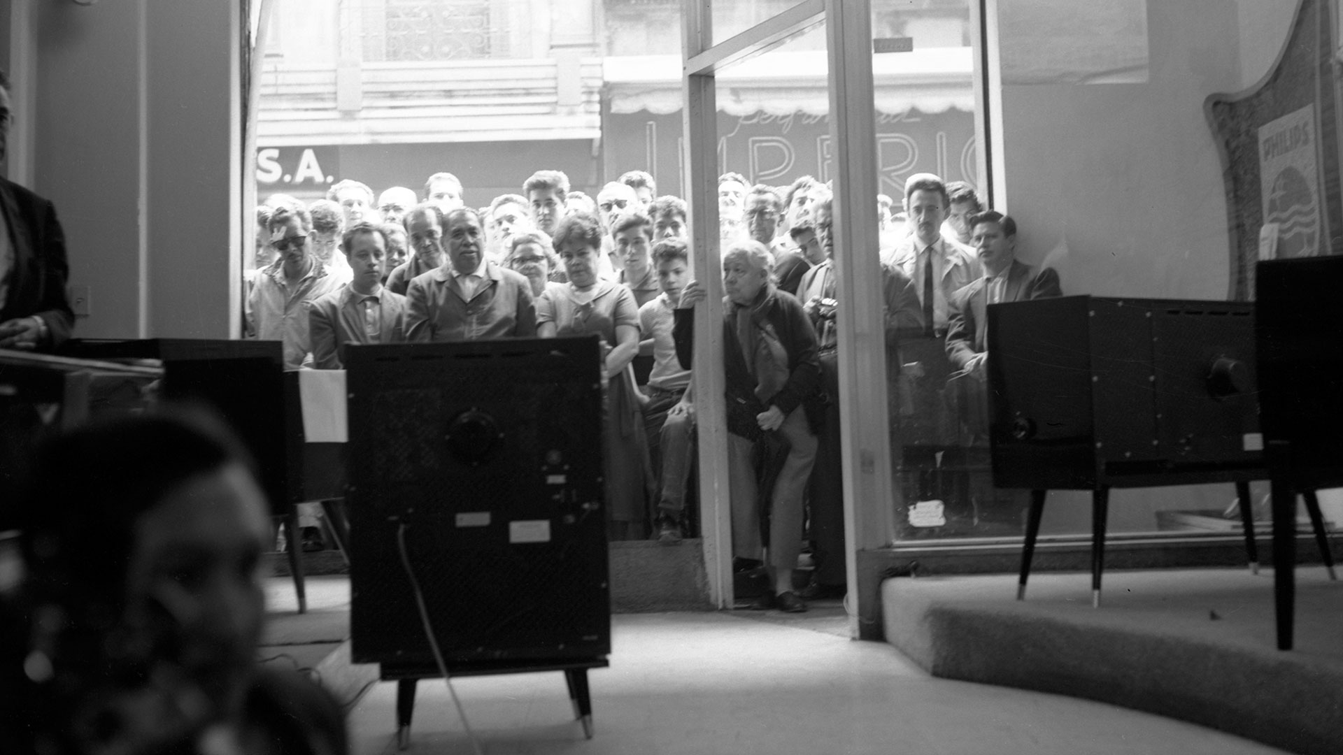 Personas asomadas al aparador de una tienda observan por televisión la visita del presidente de Estados Unidos John F. Kennedy a la Ciudad de México, junio de 1962. Colección Archivo Casasola ©19078 SC-INAH-SINAFO-Fototeca Nacional